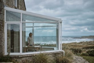Véranda en verre et aluminium blanc accolée à une maison de bord de mer, vue depuis le jardin avec vue sur le littoral