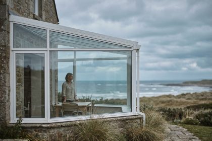 Véranda en verre et aluminium blanc accolée à une maison de bord de mer, vue depuis le jardin avec vue sur le littoral